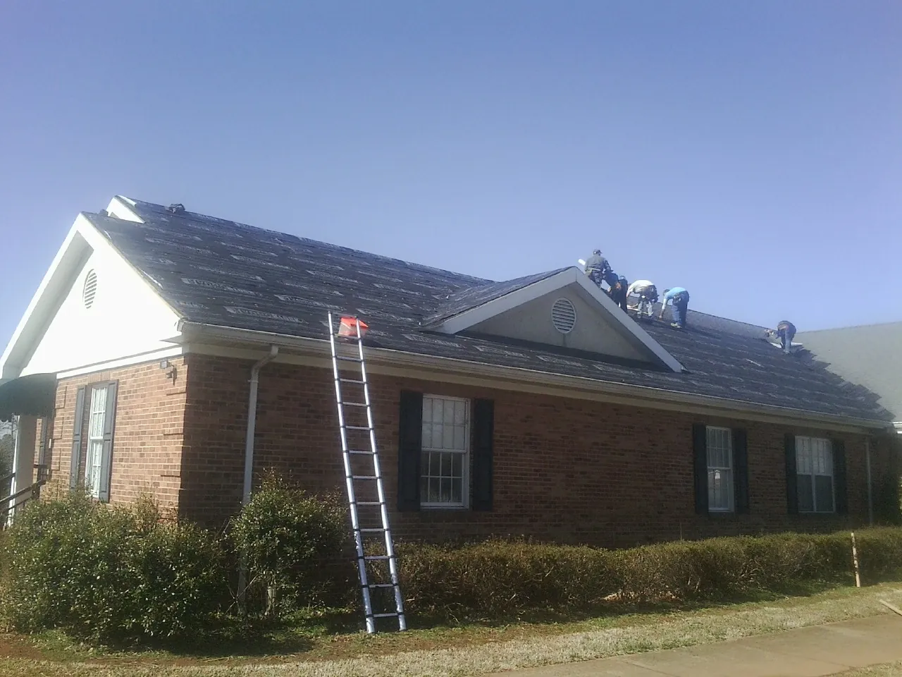 Crew tearing shingles off a brick ranch roof with a ladder set — Emergency Roof Repair work by our crew in Mayetta