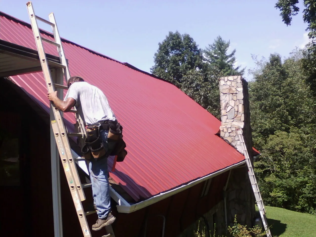 Roofer on a ladder beside a red metal roof and stone chimney — Soffit & Fascia Repair by Roofing Brothers in Mayetta